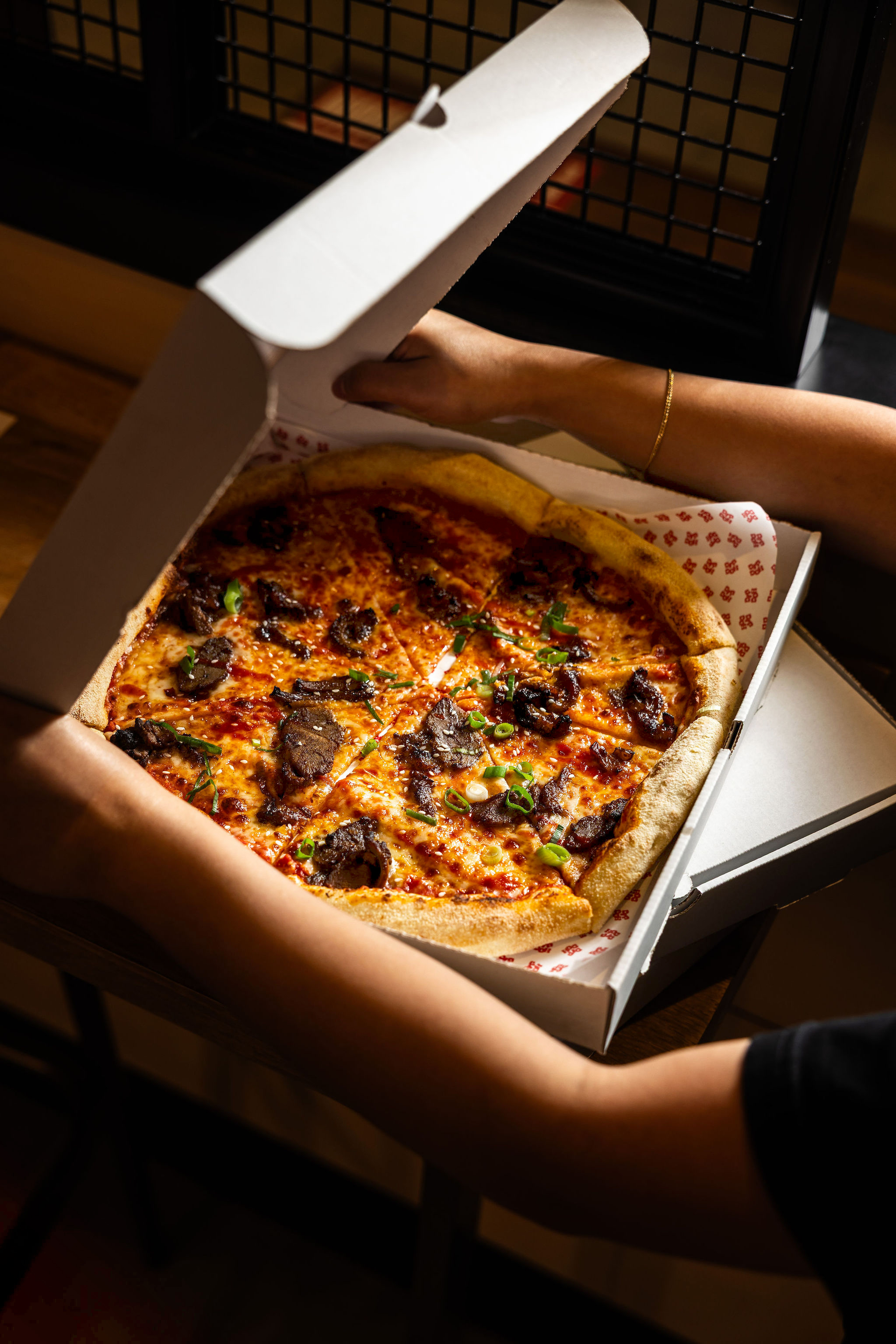 Person opening a pizza box containing a freshly baked round pizza topped with meat and spring onions.